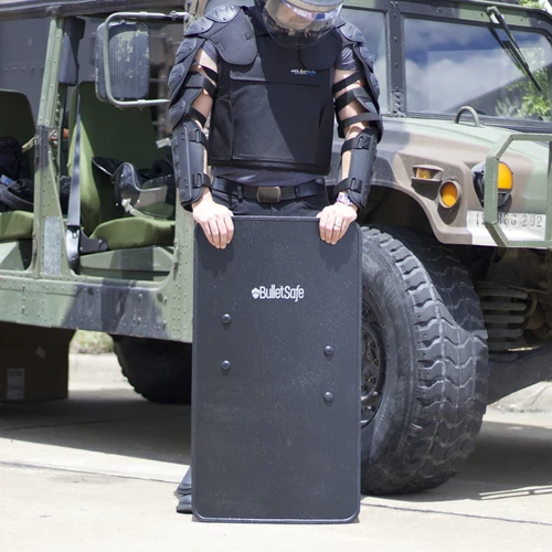 a man in full body armor holding a handgun shield while standing beside an army truck